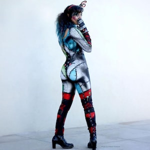 May include: A full-body jumpsuit featuring a patchwork design with black, white, blue, and red sections. The garment includes printed stitch patterns, dark red bands on the legs, and a headpiece with a black scissor icon attached to the top.