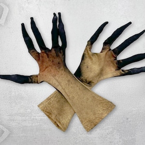 May include: A pair of costume gloves with a pale tan base and black fingertips. The gloves are crossed over each other, with the fingers spread wide. The gloves are likely made of a flexible material. The text "SHAPE SHIFTERZ" is visible in the background.