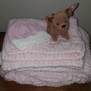 May include: A pink knitted baby blanket with a white trim and a pink knitted hat. A brown plush toy dog is sitting on top of the blanket.