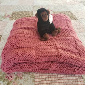 May include: A pink knitted throw blanket with a square pattern. The blanket is folded and a small black monkey plush toy is sitting on top of it.