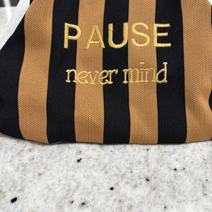 May include: A fabric pouch featuring vertical black and tan stripes. The word PAUSE is embroidered in gold thread in a sans-serif font, with the words never mind embroidered below it in a smaller, gold-colored serif font.