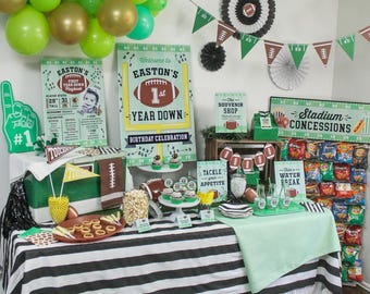 Può includere: Allestimento per una festa di compleanno a tema calcio. Decorazioni includono palloncini verdi e dorati, striscioni e cartelli con grafiche di calcio. Il tavolo è coperto da una tovaglia a righe bianche e nere e presenta snack e dolcetti.