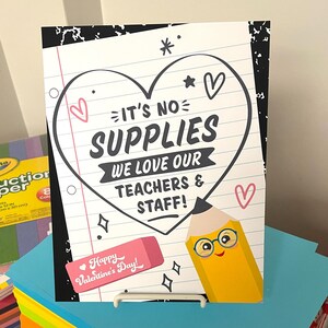 May include: A sign with a heart shape and the text "It's No Supplies We Love Our Teachers & Staff!" with a cartoon pencil with glasses and a happy face. The sign is on a stand and there are stacks of colorful paper behind it.