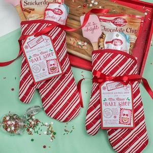 May include: Two red and white striped oven mitts with red ribbons and gift tags that say "North Pole Deliver" and "Reindeer Air Mail Drop from Mrs. Claus' Bake Shop". The tags have a red and white candy cane, a red and white striped envelope, and a red and white stamp with a picture of Mrs. Claus. The mitts are filled with baking supplies, including a box of Betty Crocker Snickerdoodle cookie mix, a jar of Betty Crocker cookie icing, a box of Betty Crocker chocolate chip cookie mix, and a pink spatula that says "all is bright".