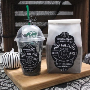 May include: Two black and white Halloween-themed drink containers. One is a clear plastic cup with a black and white label that reads "Sleepless Nights Guaranteed Night Owl Elixir Hocus Pocus Magic Infused for Focus Just Add Scream & Sugar Love, The McKowns". The other is a white paper bag with a black and white label that reads "Sleepless Nights Guaranteed Night Owl Elixir Hocus Pocus Magic Infused for Focus Just Add Scream & Sugar Love, The McKowns".