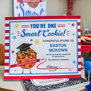 Peut inclure: Un certificat de fin d'études rouge, blanc et bleu avec un biscuit de dessin animé portant un bonnet de graduation et tenant un crayon. Le certificat indique "You're One Smart Cookie!" et "Congratulations to Easton McKown for the successful completion of preschool at The McKown Academy."