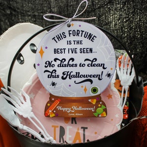 May include: A Halloween-themed gift basket with a fortune cookie-style sign that reads "This fortune is the best I've seen... No dishes to clean this Halloween!" The basket also includes paper plates, plastic forks, and a small pumpkin.