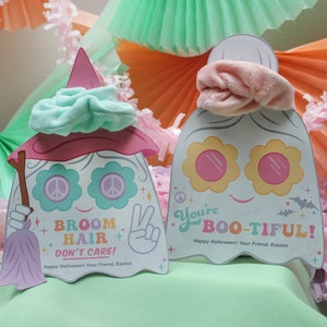 Op de afbeelding: Twee spookvormige Halloween-kaarten in roze en groen. De kaart links heeft een groen scrunchie bovenop en zegt "Broom Hair + Don't Care!" De kaart rechts heeft een roze scrunchie bovenop en zegt "You're Boo-tiful!"