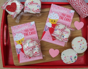 May include: Two pink Valentine's Day cards with the text "Valentine You Take The Cake!" and a cartoon cupcake. Each card is tied with red and white twine to a Hostess cupcake. Two additional Hostess cupcakes are on a red tray with pink glitter hearts.