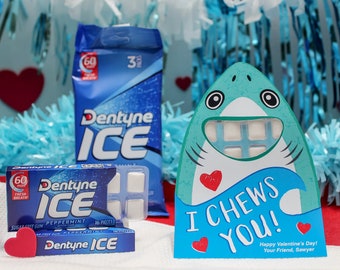 May include: A Valentine's Day card featuring a cartoon shark with a blue and green body and a red heart. The shark's mouth is open and has a pack of Dentyne Ice gum inside. The card says "I Chews You!" and "Happy Valentine's Day! Your Friend, Sawyer".