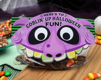 May include: A purple goblin face with big eyes and a toothy grin printed on a clear plastic bag filled with chocolate bark and candy corn. The bag has the text "Here's to Goblin' Up Halloween Fun!" printed on it.