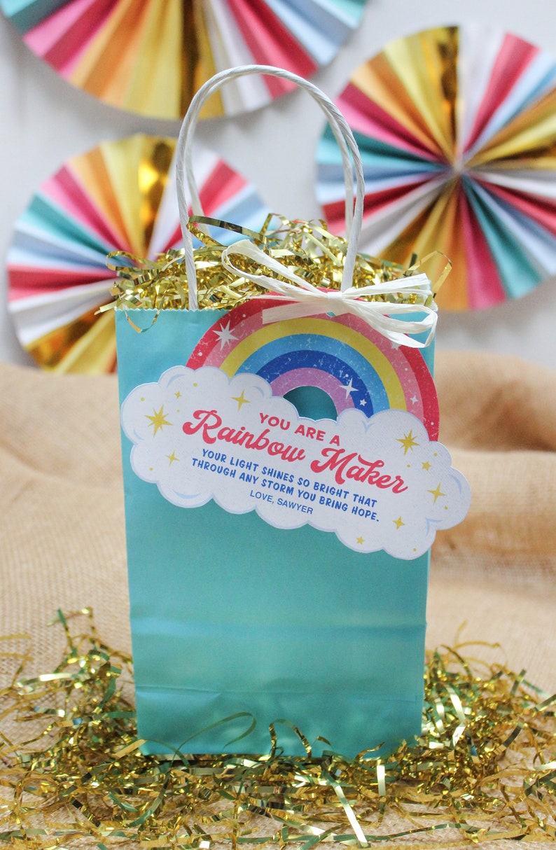 May include: A turquoise gift bag with white handles and a rainbow-themed tag that reads "You are a Rainbow Maker." The bag is filled with gold tinsel and is decorated with colorful paper fans.