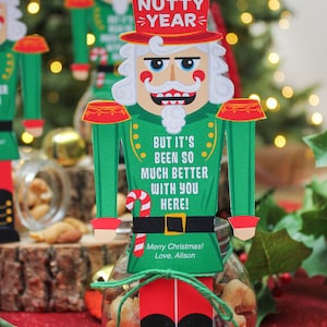 May include: A nutcracker-themed gift tag with the text "Another Nutty Year! But it's been so much better with you here! Merry Christmas! Love, Alison." The tag is attached to a glass jar filled with mixed nuts.