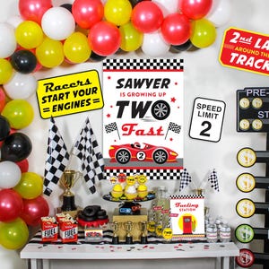 May include: A race car themed birthday party setup with balloons, signs, and decorations. The decorations include a sign that says "Racers Start Your Engines", a sign that says "Sawyer is growing up Two Fast", and a sign that says "2nd Lap Around the Track".