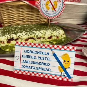 May include: A white card with red and blue checkered borders and a cartoon noodle holding a blue pencil. The card reads "Gorgonzola Cheese, Pesto, and Sun-Dried Tomato Spread".