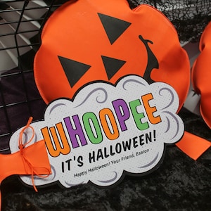 May include: A white and grey cloud-shaped sign with the text "WHOOPEE IT'S HALLOWEEN! Happy Halloween! Your Friend, Easton" in colourful letters. The sign is decorated with swirls and is attached to an orange pumpkin-shaped cushion.