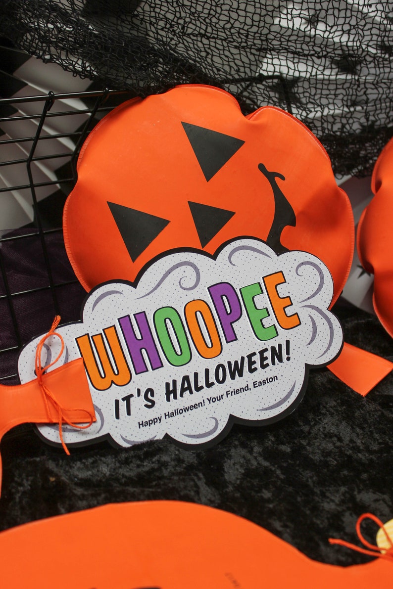May include: A white and grey cloud-shaped sign with the text "WHOOPEE IT'S HALLOWEEN! Happy Halloween! Your Friend, Easton" in colourful letters. The sign is decorated with swirls and is attached to an orange pumpkin-shaped cushion.
