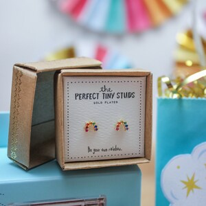 May include: Gold-plated stud earrings in a gift box. The earrings are shaped like rainbows with colorful gemstones. The box reads "the PERFECT TINY STUDS" and "Be your own rainbow."