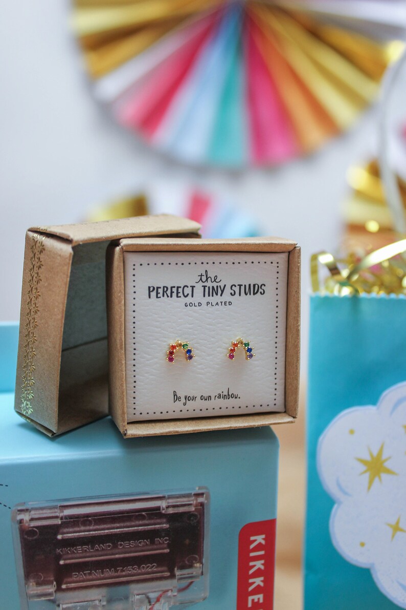 May include: Gold-plated stud earrings in a gift box. The earrings are shaped like rainbows with colorful gemstones. The box reads "the PERFECT TINY STUDS" and "Be your own rainbow."