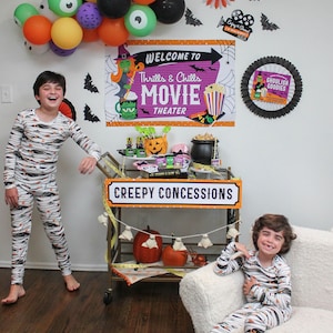 May include: Two children dressed in black and white striped pajamas with a Halloween theme stand in front of a Halloween movie night themed party set up. The party decorations include a banner that says "Welcome to Thrills & Chills Movie Theater", a black and white fan with a witch on it, and a black and white fan with the words "Ghoulish Goodies" on it. There is a cart with the words "Creepy Concessions" on it. The cart is decorated with orange pumpkins, white ghosts, and black bats.
