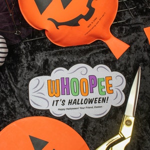 May include: Two orange pumpkin-shaped whoopie cushions with black jack-o'-lantern faces. A pair of gold scissors and a white card with the text "WHOOPEE IT'S HALLOWEEN! Happy Halloween! Your Friend, Easton" are on a black velvet surface.