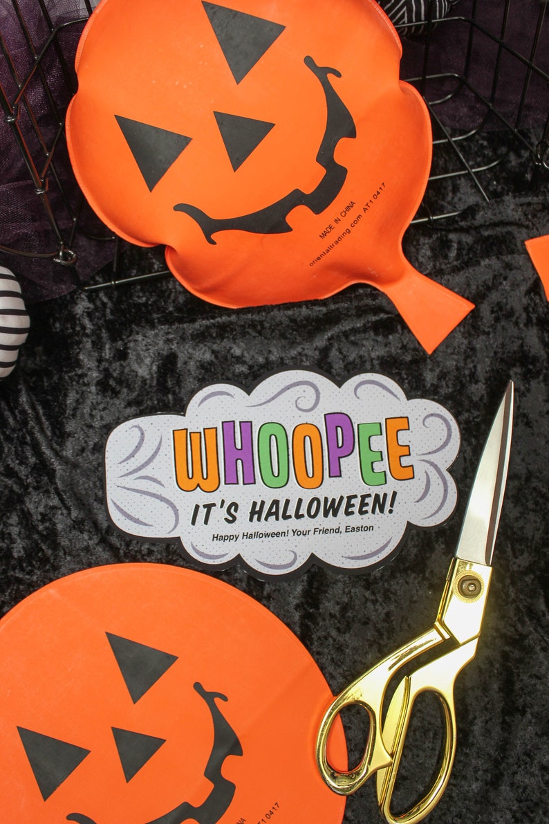 May include: Two orange pumpkin-shaped whoopie cushions with black jack-o'-lantern faces. A pair of gold scissors and a white card with the text "WHOOPEE IT'S HALLOWEEN! Happy Halloween! Your Friend, Easton" are on a black velvet surface.