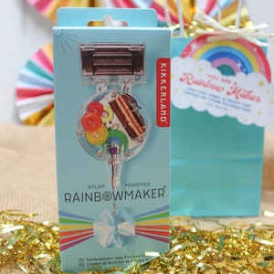 May include: A light blue box with a solar-powered Rainbowmaker toy. The box features a clear plastic window showing the internal gears and components. A gift bag with a rainbow tag and gold tinsel complete the scene.