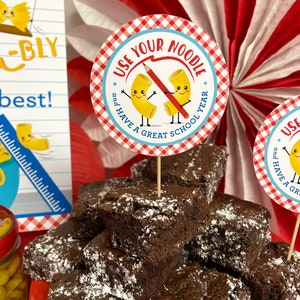 May include: A stack of chocolate brownies topped with powdered sugar, with a red and white checkered paper circle with the text "Use Your Noodle and Have a Great School Year" on a toothpick in the center.