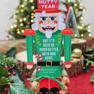 May include: A nutcracker figurine holding a jar of mixed nuts. The nutcracker is wearing a red hat with the words "Another Nutty Year" on it. The jar is tied with a green ribbon and has a tag that says "Merry Christmas! Love, Alison".