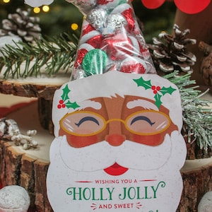 May include: A white Santa Claus face with a red nose and a green holly sprig on the side. The Santa face is holding a clear plastic bag filled with red and silver foil-wrapped chocolates. The bag is tied with a white ribbon. The text on the Santa face reads "Wishing you a Holly Jolly and Sweet Christmas! Love, Sawyer and Easton".