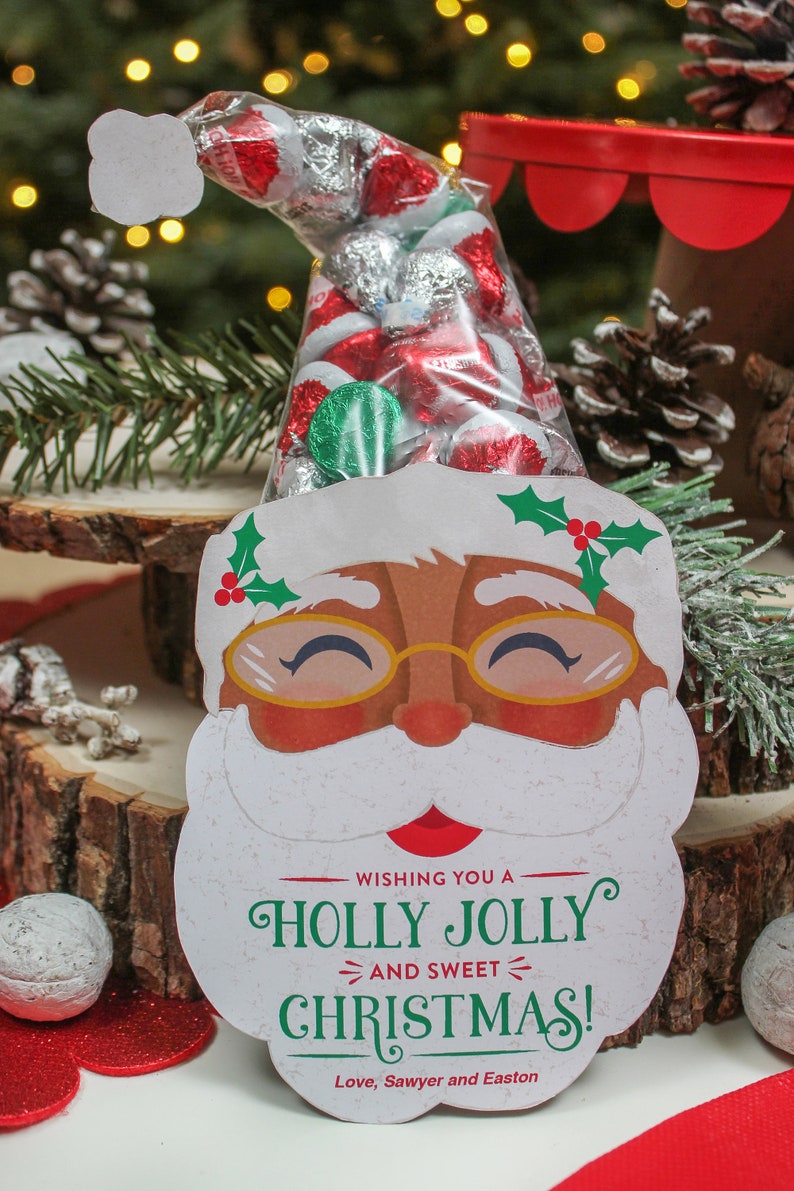 May include: A white Santa Claus face with a red nose and a green holly sprig on the side. The Santa face is holding a clear plastic bag filled with red and silver foil-wrapped chocolates. The bag is tied with a white ribbon. The text on the Santa face reads "Wishing you a Holly Jolly and Sweet Christmas! Love, Sawyer and Easton".