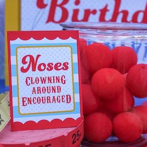 May include: Red and blue sign with white polka dots that reads "Noses Clowning Around Encouraged". A glass jar filled with red foam balls sits next to the sign. A stack of red and yellow tickets with the number 25&cent; printed on them are in front of the sign.