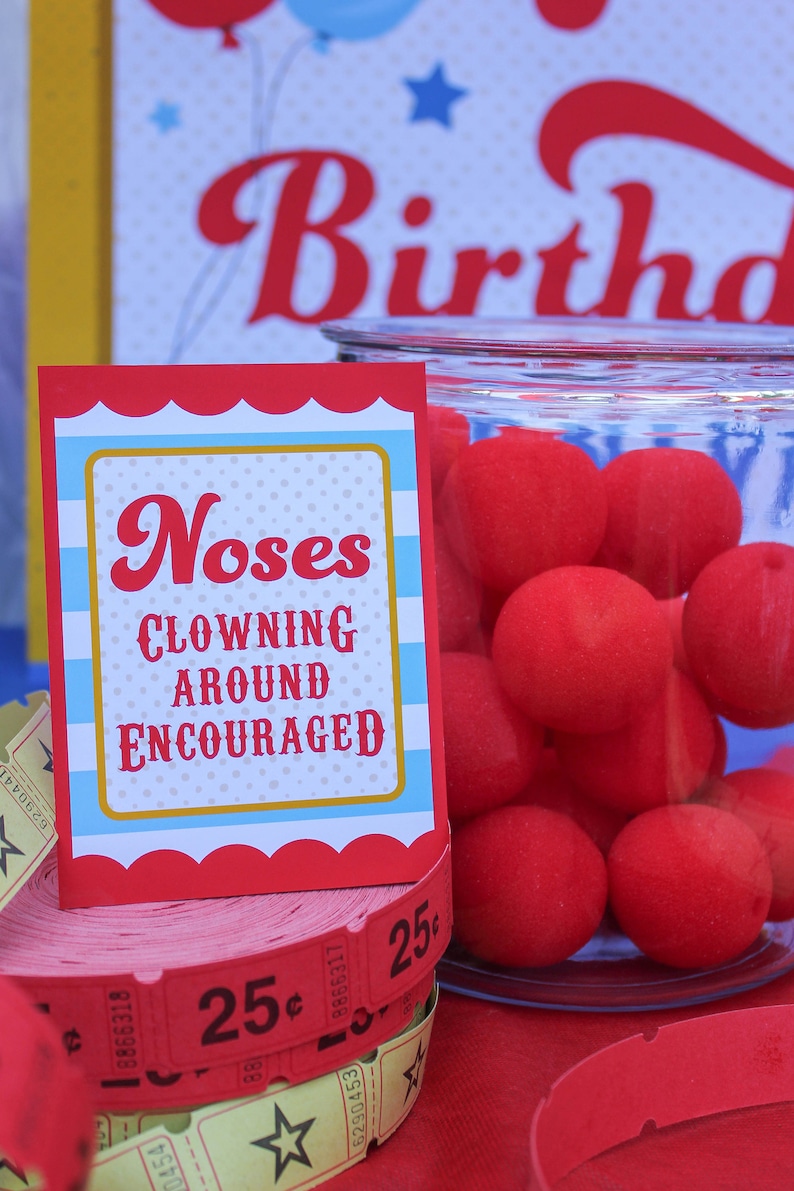 May include: Red and blue sign with white polka dots that reads "Noses Clowning Around Encouraged". A glass jar filled with red foam balls sits next to the sign. A stack of red and yellow tickets with the number 25&cent; printed on them are in front of the sign.