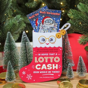 Pode incluir: Uma meia vermelha com um rosto branco e &oacute;culos, segurando dois bilhetes de loteria. A meia tem o texto "Na esperan&ccedil;a de que um Lotto Cash chegue em breve!" e est&aacute; cercada por pequenas &aacute;rvores de Natal artificiais e moedas de ouro.
