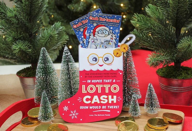 Pode incluir: Uma meia vermelha com um rosto branco e &oacute;culos, segurando dois bilhetes de loteria. A meia tem o texto "Na esperan&ccedil;a de que um Lotto Cash chegue em breve!" e est&aacute; cercada por pequenas &aacute;rvores de Natal artificiais e moedas de ouro.