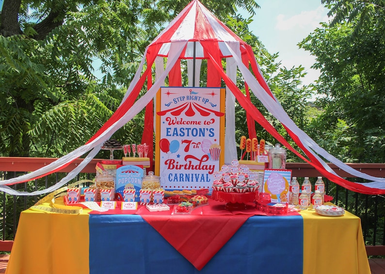 Peut inclure: Une d&eacute;coration de f&ecirc;te d'anniversaire sur le th&egrave;me du carnaval avec un code couleur rouge, blanc et bleu. Une pancarte indique "Bienvenue au 7e anniversaire de Easton". La table est remplie de friandises, de pop-corn et de petits g&acirc;teaux.