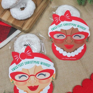May include: Two clear plastic bags filled with powdered doughnuts and decorated with red and white Christmas-themed gift tags. The tags feature a cartoon woman wearing red glasses and a red bow. The text on the tags reads "Sweetest Christmas Wishes!" and "Love, Sawyer and Easton".