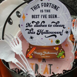 May include: A black cauldron filled with Halloween treats, including a pink plate with the words "Trick or Treat" and a white paper plate with a ghost face. There is a white plastic skeleton fork and a fortune cookie with the text "This fortune is the best I've seen... No dishes to clean this Halloween! Happy Halloween! Love, Alison".