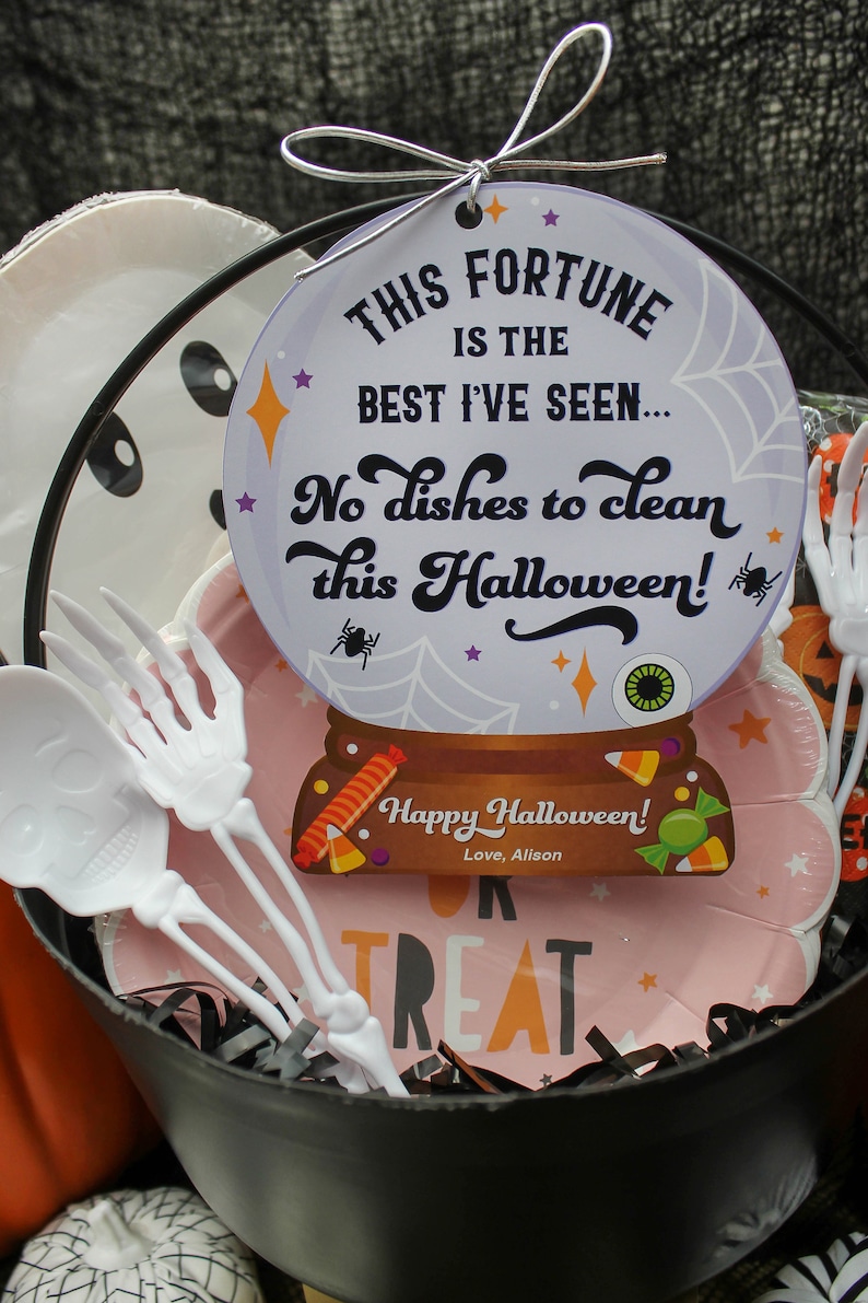 May include: A black cauldron filled with Halloween treats, including a pink plate with the words "Trick or Treat" and a white paper plate with a ghost face. There is a white plastic skeleton fork and a fortune cookie with the text "This fortune is the best I've seen... No dishes to clean this Halloween! Happy Halloween! Love, Alison".