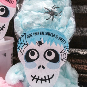 Puede incluir: Algod&oacute;n de az&uacute;car azul en una bolsa de pl&aacute;stico transparente con una pegatina de calavera blanca que dice "Espero que tu Halloween sea dulce. Con cari&ntilde;o, Sawyer & Easton". Hay ara&ntilde;as negras en el algod&oacute;n de az&uacute;car y la pegatina.