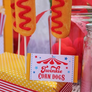 May include: A yellow and white polka dot box with a red and white striped ribbon. The box is filled with Twinkie corn dogs, which are yellow cake pops with red frosting. A sign reads "Twinkie Corn Dogs" with a red and white circus tent graphic.
