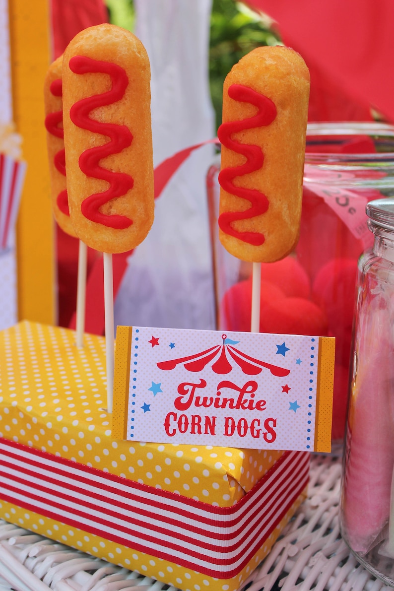 May include: A yellow and white polka dot box with a red and white striped ribbon. The box is filled with Twinkie corn dogs, which are yellow cake pops with red frosting. A sign reads "Twinkie Corn Dogs" with a red and white circus tent graphic.