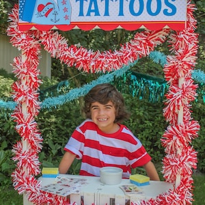 May include: A young boy wearing a red and white striped shirt stands behind a white wooden stand decorated with red and white pom-poms. The stand has a sign that reads "Easton's Carnival Tattoos".