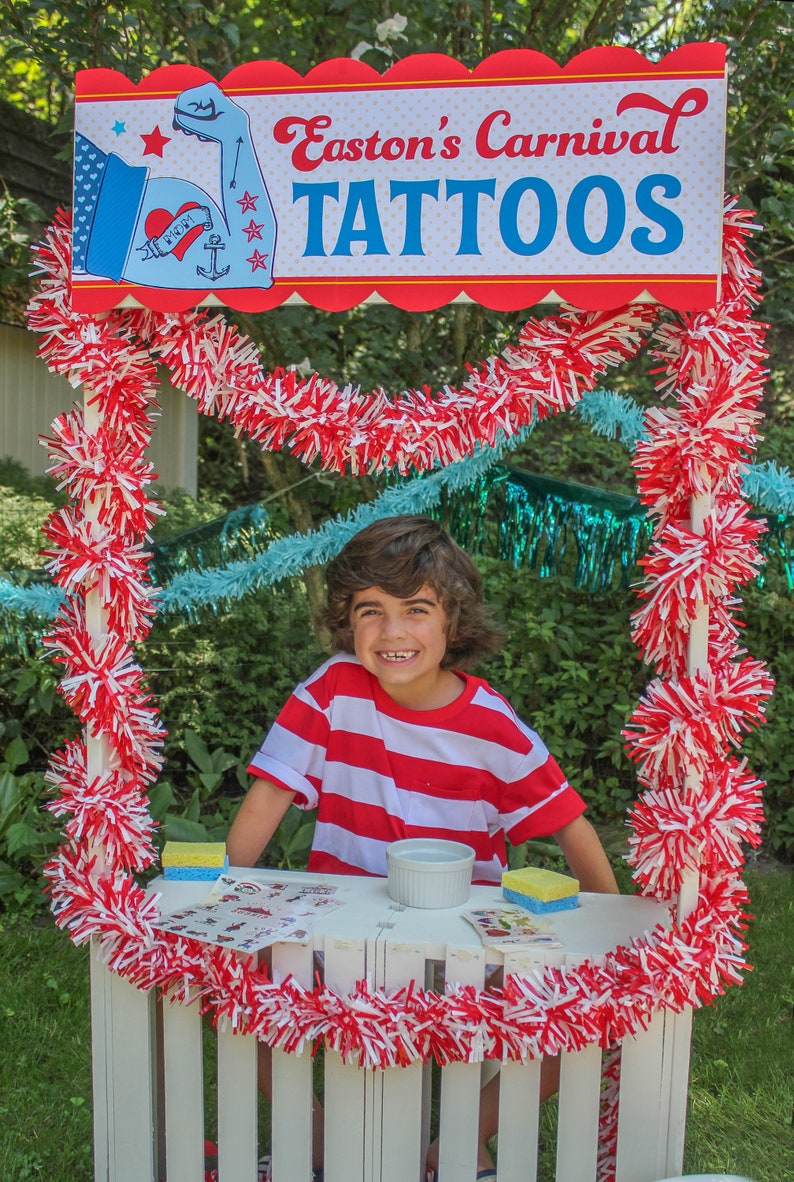May include: A young boy wearing a red and white striped shirt stands behind a white wooden stand decorated with red and white pom-poms. The stand has a sign that reads "Easton's Carnival Tattoos".