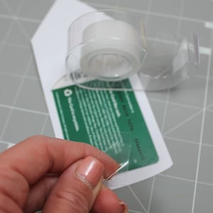 May include: A clear tape dispenser with a roll of transparent tape is positioned over a white card with green text. A hand is pulling a piece of tape from the dispenser. The background is a gray cutting mat.