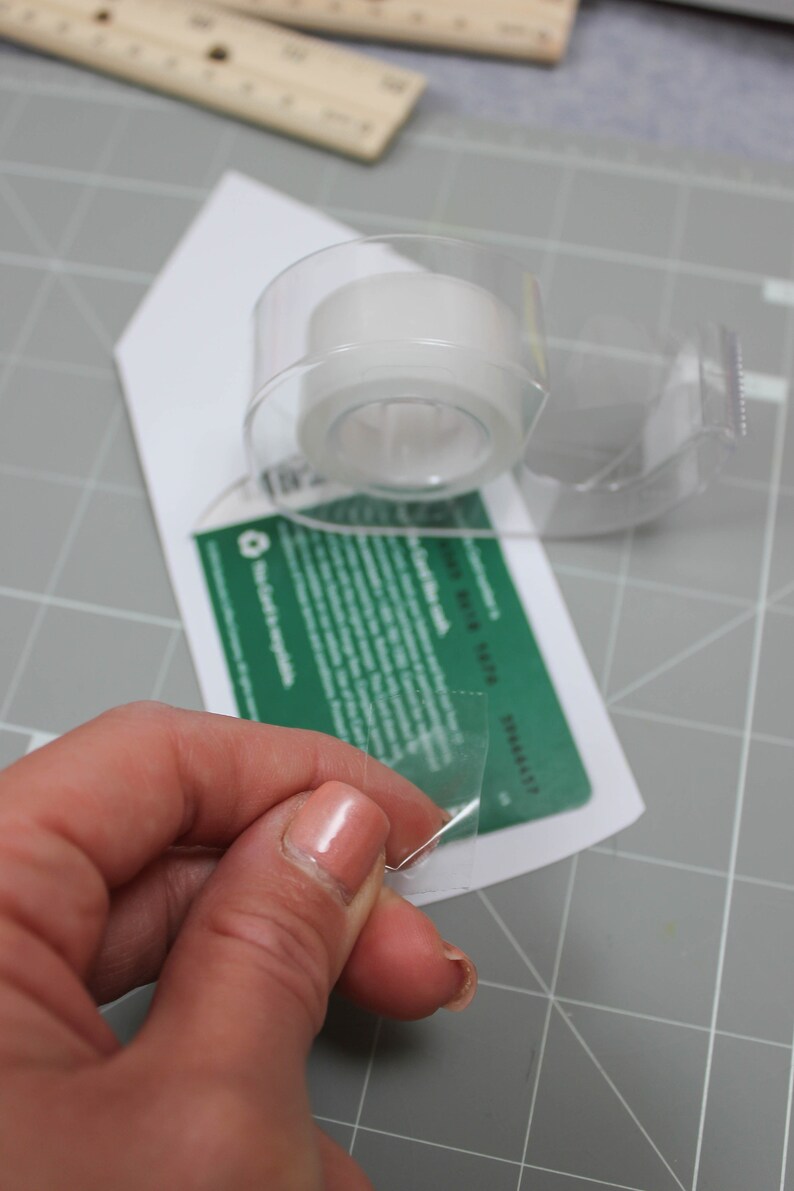May include: A clear tape dispenser with a roll of transparent tape is positioned over a white card with green text. A hand is pulling a piece of tape from the dispenser. The background is a gray cutting mat.