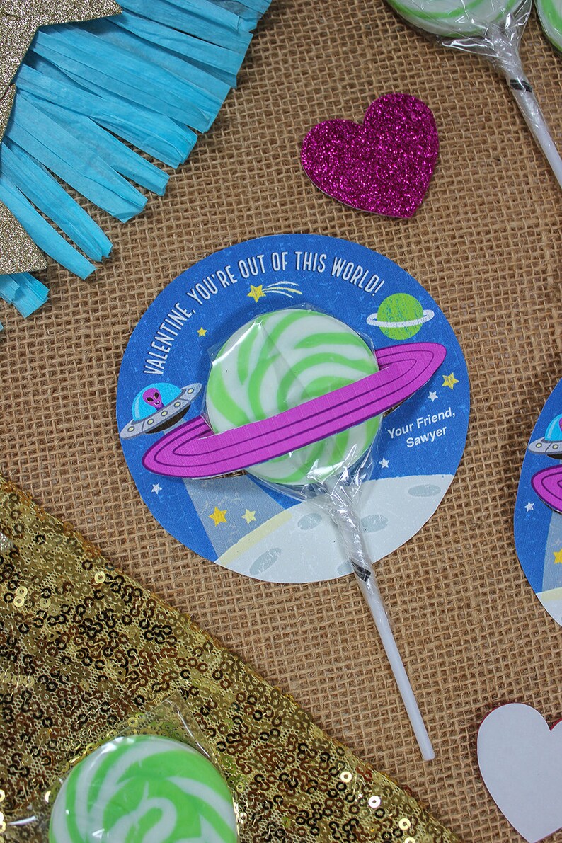 Op de afbeelding: Een groene en witte swirl lollipop met een roze en paarse ring eromheen. De lollipop is bevestigd aan een stok en bevindt zich op een blauwe en witte cirkel met de tekst "Valentine, you're out of this world!" en "Your Friend, Sawyer".