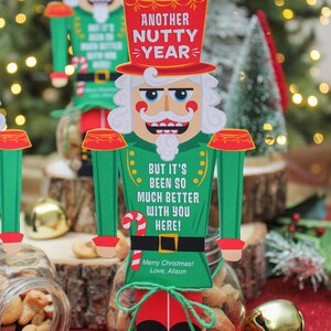 May include: A nutcracker figurine with a red hat and green uniform holds a jar of mixed nuts. The figurine has a sign that reads "Another Nutty Year! But it's been so much better with you here!" and "Merry Christmas! Love, Alison".