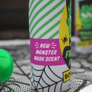 May include: A green and yellow can with a white and green striped background. The can has a pink label that says "NEW MONSTER MUSK SCENT" and a black and white spider graphic. The can is sitting on a black spiderweb-patterned surface.