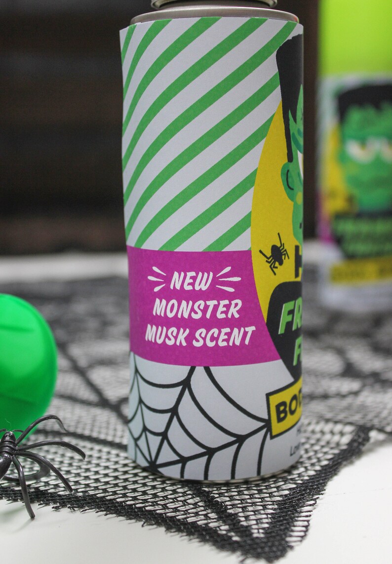 May include: A green and yellow can with a white and green striped background. The can has a pink label that says "NEW MONSTER MUSK SCENT" and a black and white spider graphic. The can is sitting on a black spiderweb-patterned surface.